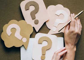 Hands holding pen next to paper question marks on notebook, symbolizing inquiry and creativity.